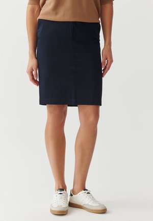 Femme portant une jupe bleu marine foncé longueur genou, un haut marron et des baskets blanches avec semelles en gomme, debout sur un fond blanc.