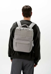 Zaino in tessuto grigio con spallacci imbottiti e maniglia superiore; presenta una tasca frontale con zipper e tasche laterali. Design minimalista.