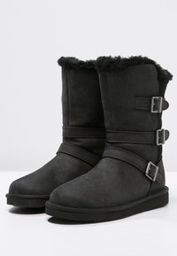 Bottes cheville en daim noir avec bordure en fourrure, dotées de deux brides ajustables et de boucles en métal sur le côté. Semelles plates en caoutchouc.