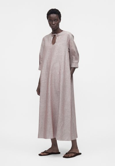 Γυναίκα που φοράει ένα φαρδύ, ρόζ φόρεμα με ρίγες μέχρι τους αστραγάλους, με μανίκια τριών τετάρτων και μικρό δετό άνοιγμα στο λαιμό, συνδυασμένο με μαύρα σαγιονάρια.