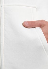 Hvid, struktureret stof af en sweatshirt med lynlås, med en synlig lomme og en hånd placeret mod den, der fremhæver dens funktionelle design.