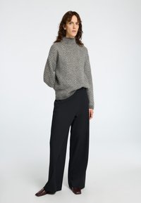 Grayer Strickpullover im Fischgrätmuster mit hohem Kragen, kombiniert mit schwarzen weit geschnittenen Hosen und spitzen bordeauxroten Schuhen.