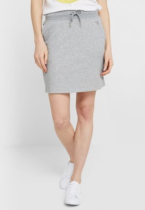 Femme portant une jupe gris mi-longue à cordon avec poches, un t-shirt blanc et des baskets blanches, debout jambes croisées.