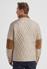 Gesteppte beige Jacke mit braunen Cordakzenten an den Ellbogen, ausgestattet mit einem klassischen Kragen und Druckknöpfen an den Manschetten.