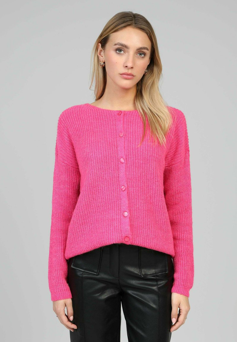Cardigan en maille rose vif avec un col rond, des manches longues et des boutons sur le devant. Caractérisé par une coupe ample et une texture côtelée. Associé à un pantalon noir.