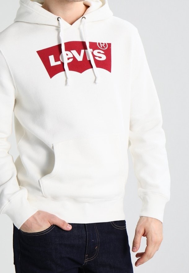 Homme portant un sweat à capuche blanc avec un logo Levi's rouge sur la poitrine et un jean bleu foncé, une main dans la poche.