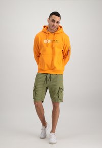 Haut à capuche orange avec logo, doté d'une capuche avec cordon de serrage ; associé à un short cargo vert avec poches latérales ; baskets blanches.