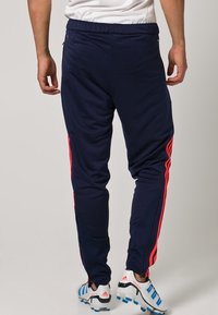 Tmavě modré sportovní kalhoty s červenými pruhy na bocích, vyrobené z hladké látky. Mají elastický pas a zúžený střih; nošeny s kopačkami.