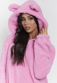 Felpa rosa, pelosa, con cappuccio e orecchie da orso, con chiusura a zip e maniche lunghe. Texture morbida con un aspetto peloso e accogliente.