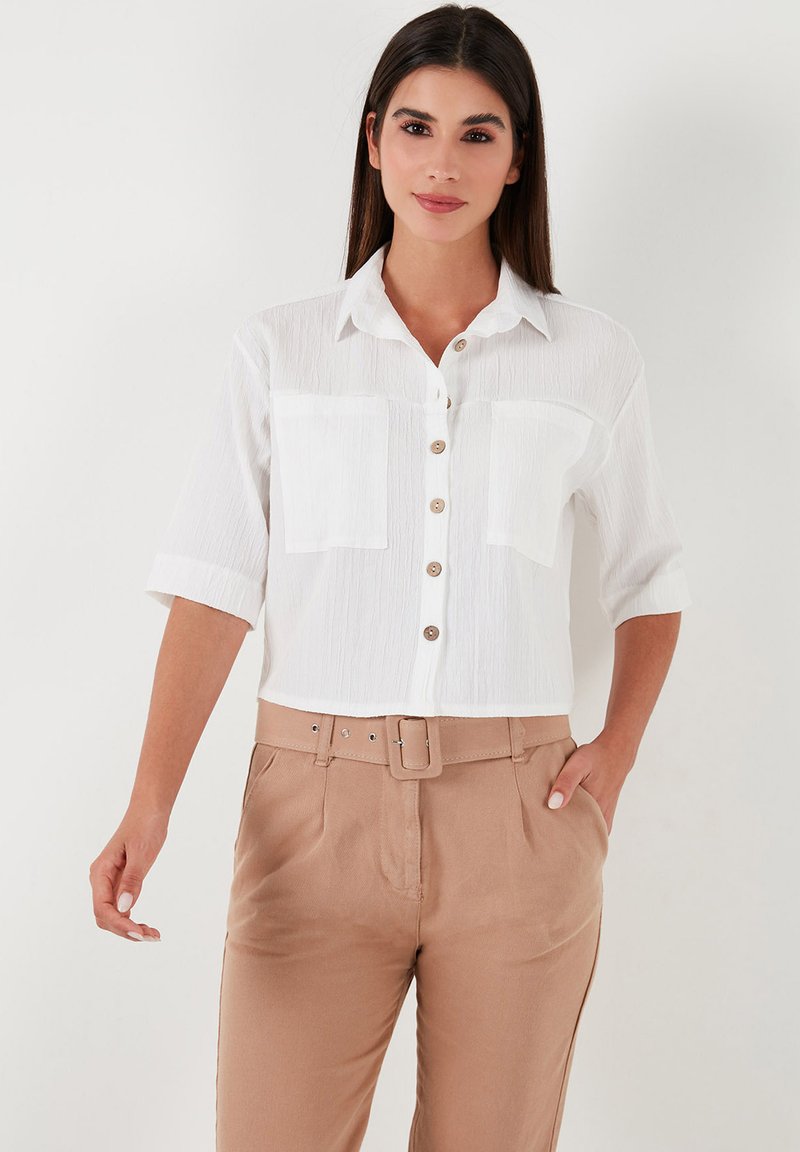 White cropped button-up shirt with short sleeves, textured fabric, two chest pockets, and beige high-waisted pants with a belt.