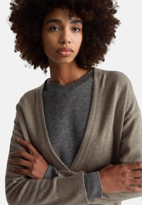 Młoda osoba z kręconymi włosami, ubrana w szary sweter i beżowy kardigan, z założonymi ramionami, o neutralnym wyrazie twarzy, na białym tle.