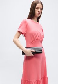 Γυναίκα με μακριά ίσια μαλλιά, φορώντας ένα κοραλί ροζ φόρεμα, κρατώντας μια μαύρη τσάντα χειρός πάνω σε ένα απλό φωτεινό φόντο.