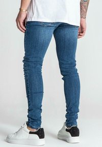 Skinny blue denim jeans with a faded wash, contrast stitching, and a back pocket detail. Paired with white sneakers featuring black accents.