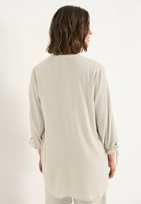 Vrouw met schouderlang bruin haar, gekleed in een losse, lichtbeige lange mouw top met gathered cuffs en strikjes, met de rug naar de camera gekeerd.