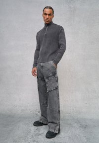 Suéter gris acanalado con media cremallera combinado con pantalones grises de estilo cargo que cuentan con múltiples bolsillos y un dobladillo deshilachado. Unos zapatos negros de deslizar completan el look.