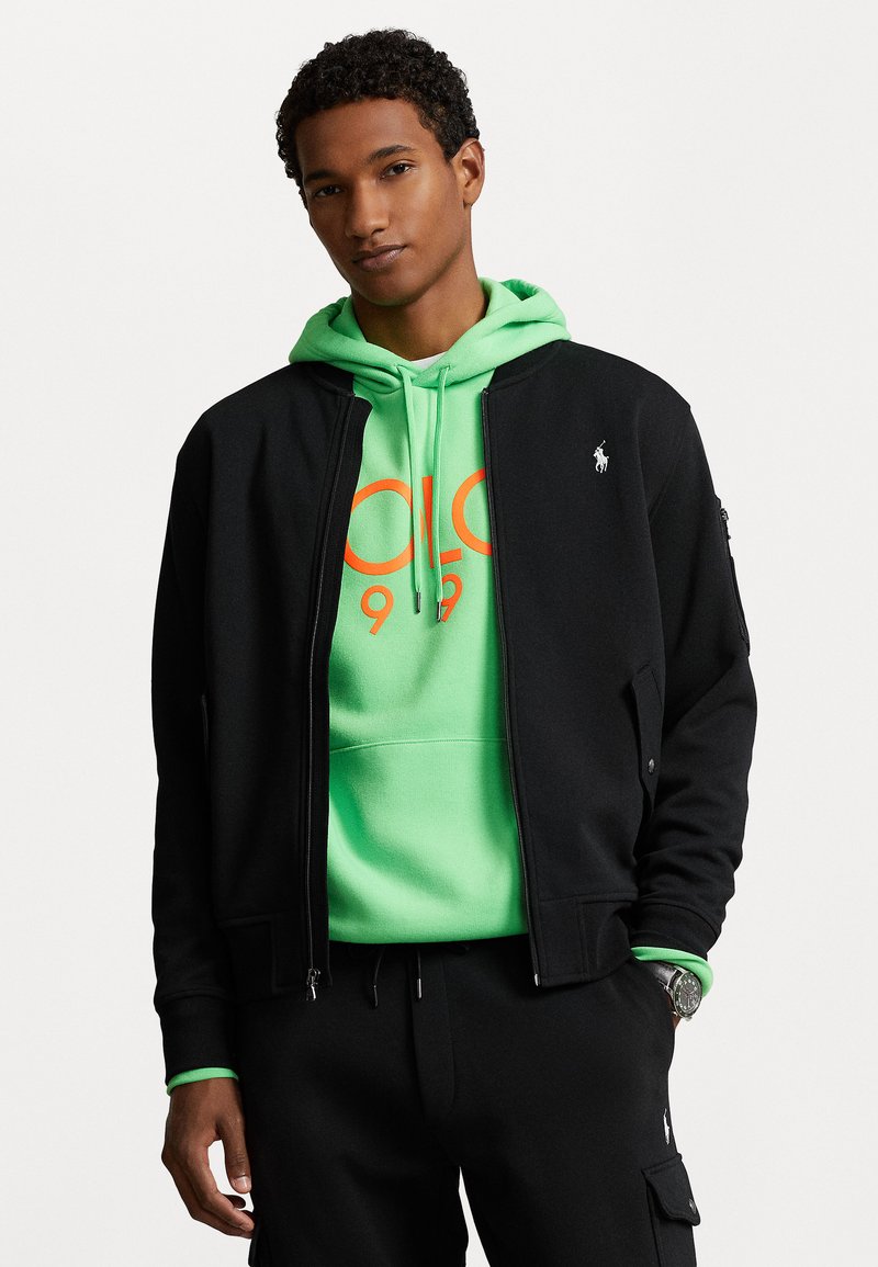 Sort bomberjakke over en lys grøn hoodie med orange grafik. Glat stof, lynlåslommer, ribbet manchetter og logo-detaljer.