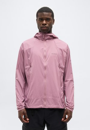 Mann trägt eine hellviolette Kapuzenjacke mit Reißverschluss und seitlichen Reißverschlusstaschen, steht vor einem schlichten weißen Hintergrund.