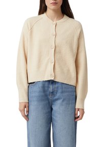 Kremowy, krótki kardigan wykonany z teksturowanej tkaniny, z okrągłym dekoltem, zapinany na guziki i z prążkowanymi mankietami. Noszony na niebieskich dżinsach.