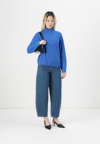Maglione blu a maglia con colletto alto, abbinato a jeans di denim a vestibilità ampia e tacchi slingback neri. Il modello tiene in mano una piccola borsa nera.