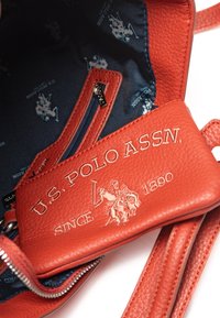 Rød læderhåndtaske med tekstureret overflade, der har en indvendig marineblå foring med brandlogo og en lynlåslomme.