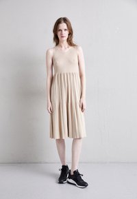 Béžové šaty bez rukávů s žebrovanou texturou a vrstvenou sukní, kombinované s černými sportovními teniskami s bílými podrážkami a tkaničkami.
