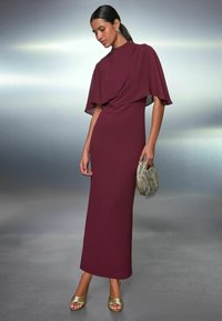 Bordeaux lang kjole med korte flagrende ærmer, tætsiddende silhuet, fremstillet af glat stof. Parret med en struktureret gylden clutch og sandaler.