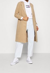 Beige lång kappa med vågade slag, bärs över en vit hoodie, kombinerad med ljusblå jeans och vita högskaftade sneakers.