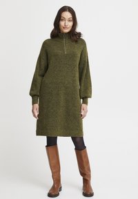 Abito verde lavorato a maglia con un alto colletto, chiusura con zip e maniche a palloncino lunghe, abbinato a stivali marroni in pelle alti fino al ginocchio. Texture liscia.