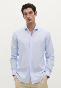 Camisa de color azul claro, de manga larga, con collar puntiagudo, botones de tela y una textura sutil. Usada por fuera, combinada con pantalones beiges.