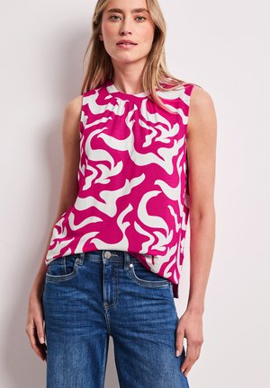 Femme portant un haut sans manches à motifs roses et blancs, rentré dans un jean en denim bleu, se tenant devant un fond gris clair.