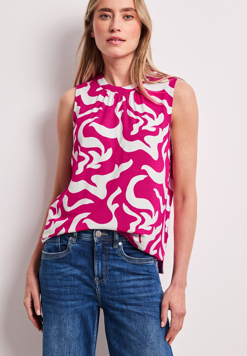 Donna che indossa una canotta senza maniche rosa con motivo bianco infilata in un paio di jeans di denim blu, in piedi contro uno sfondo grigio chiaro.