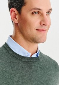 Szary dzianinowy sweter na jasnoniebieskiej koszuli z kołnierzykiem. Skupienie na czystych liniach, okrągłym dekolcie i gładkiej teksturze.