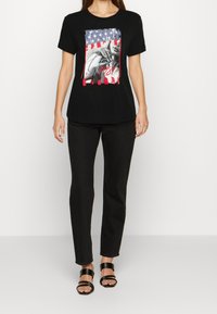 Sort bomulds t-shirt med en grafik, der viser et slidt amerikansk flag og tekst. Sættet er parret med sorte jeans og sorte remsandaler.