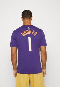 Lilla NBA-trøje med "BOOKER" i orange bogstaver og nummer "1" i hvid tekstureret skrift. Standard rund halsudskæring, korte ærmer.