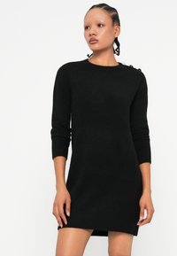 Vestido de malha preto com mangas longas, decote redondo e detalhe de botão em um ombro, apresentando uma silhueta ajustada e textura suave.