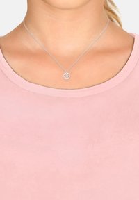Collana d'argento con un pendente circolare che presenta un design floreale, appoggiata su una camicia rosa con scollo rotondo. La catena è delicata e liscia.