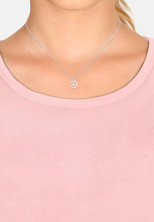 Collana d'argento con un pendente circolare che presenta un design floreale, appoggiata su una camicia rosa con scollo rotondo. La catena è delicata e liscia.