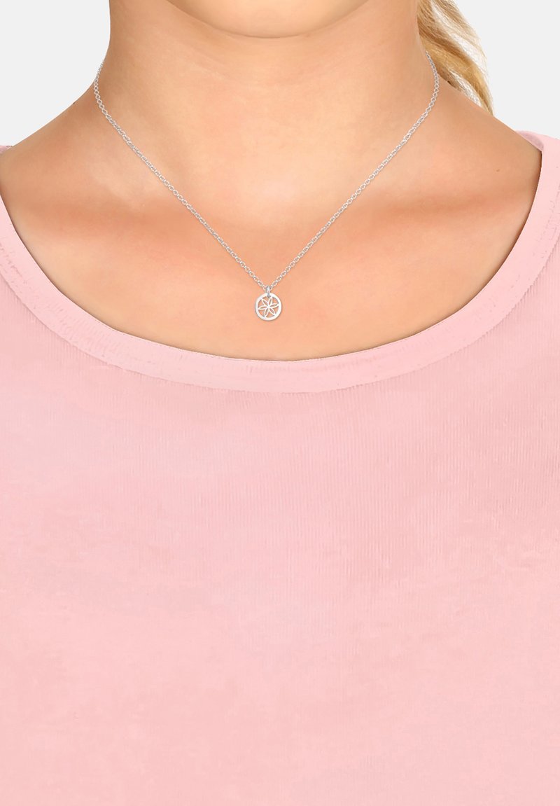 Collana d'argento con un pendente circolare che presenta un design floreale, appoggiata su una camicia rosa con scollo rotondo. La catena è delicata e liscia.