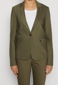 Marynarka w kolorze oliwkowej zieleni wykonana z gładkiego, lekkiego materiału. Posiada pojedynczy czarny guzik, wycięte klapy oraz dwie przednie kieszenie.