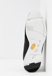 Sula av en svart och vit sneakers med orange Vibram-logotyp, placerad upprätt mot en vit hörnvägg.