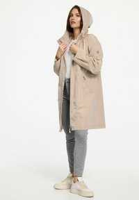 Beige waterdichte regenjas met een capuchon, frontale ritssluiting en zijzakken. Soepele textuur, boven de knie in lengte, gedragen over een witte top en grijze jeans.