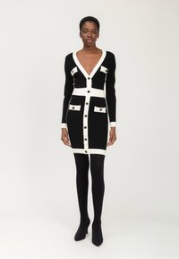 Schwarzes Strickkleid mit weißen Akzenten, V-Ausschnitt, langen Ärmeln, durchgehender Knopfleiste, zwei Brusttaschen und taillierter Passform. Kombiniert mit schwarzen Strumpfhosen.
