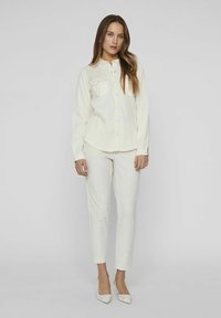 Camicia bianca con bottoni, maniche lunghe e taschino sul petto, abbinata a pantaloni crema a sigaretta e décolleté a punta. Tessuto morbido e leggero.