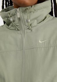 Svijetlozelena jakna na zip s visokim ovratnikom, podešavanjima na vezicu i malim bež Nike logotipom na prsima. Glatka, meka tkanina.