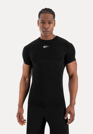 Uomo atletico con capelli intrecciati che indossa una maglietta nera aderente a maniche corte con un logo bianco, in piedi davanti a uno sfondo chiaro e semplice.