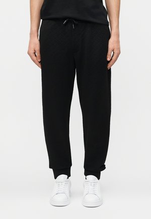 Mees, kes kannab musta tekstuuriga dressipükse, millel on nöörvöökoht, ja valgeid tossusid, seisab ühevärvilise heleda tausta ees.