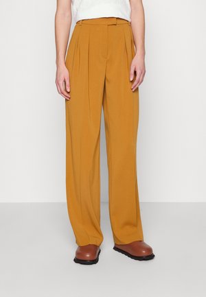 Pantalons à jambes larges couleur jaune moutarde, avec un devant plissé, une taille haute et une finition en tissu lisse. Des chaussures marron complètent le look.