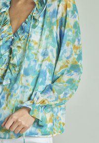 Leichte Bluse mit langen Ärmeln, rüschigem Ausschnitt und Knopfdetails. Sie hat ein abstraktes Muster in Blau, Grün und Gelb. Weicher Stoff.
