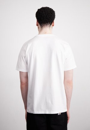 Man met kort krullend haar, een effen wit t-shirt met korte mouwen en een zwarte broek, staand met zijn rug naar voren tegen een effen achtergrond.