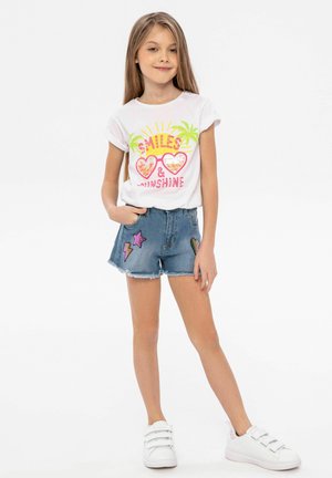 Vit t-shirt med färggrafisk text och palmträd, i kombination med denimshorts med stjärn- och blixtapplikationer, vita sneakers.